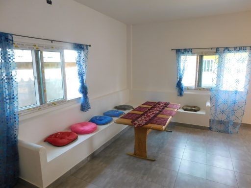 A bright room with a table, colorful cushions, and windows covered with light curtains.